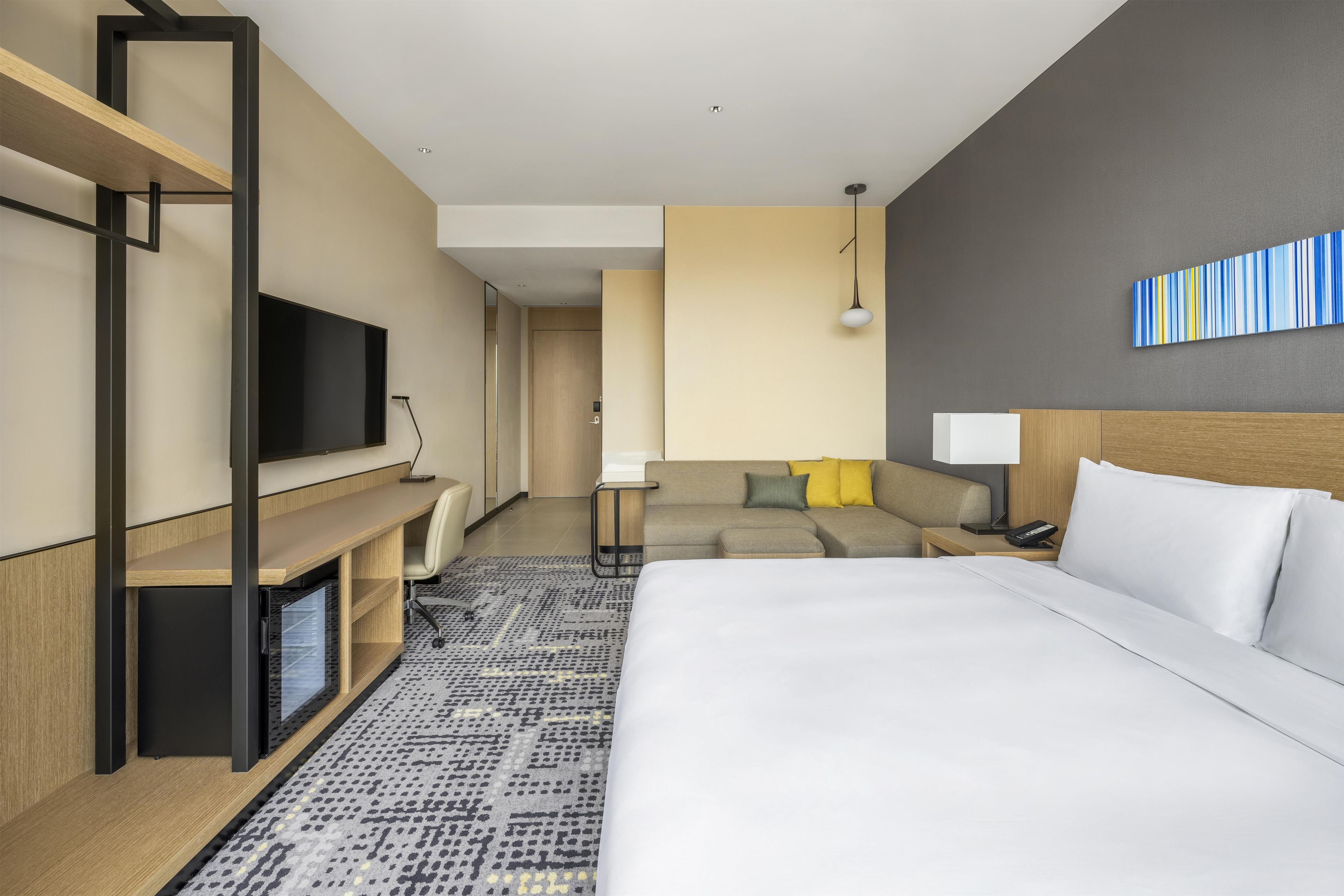 A modern hotel room with a large bed, a desk with a flat-screen TV, a sofa, and a patterned rug.