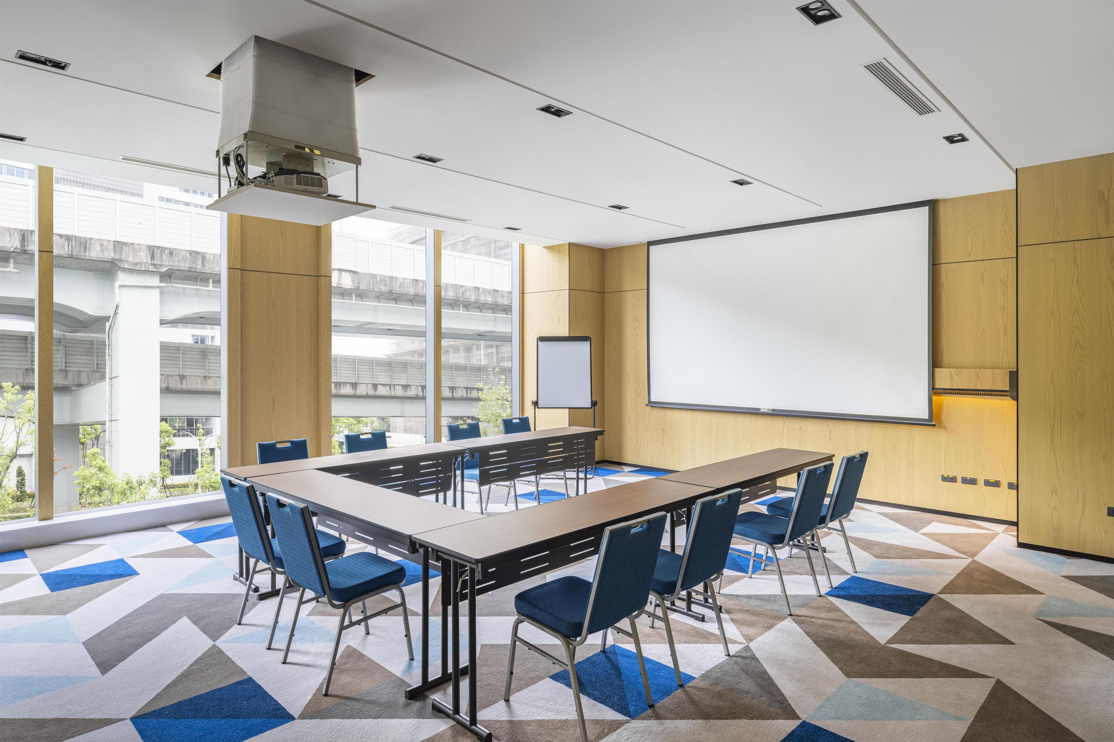 Une salle de conférence équipée d’une grande table, de chaises, d’un écran de projection et offrant une vue sur un espace extérieur.
