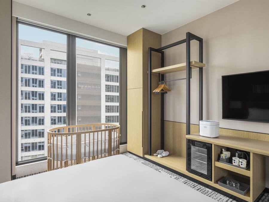 A modern hotel room with a large window, a crib, a TV, and a wooden console with shelves.