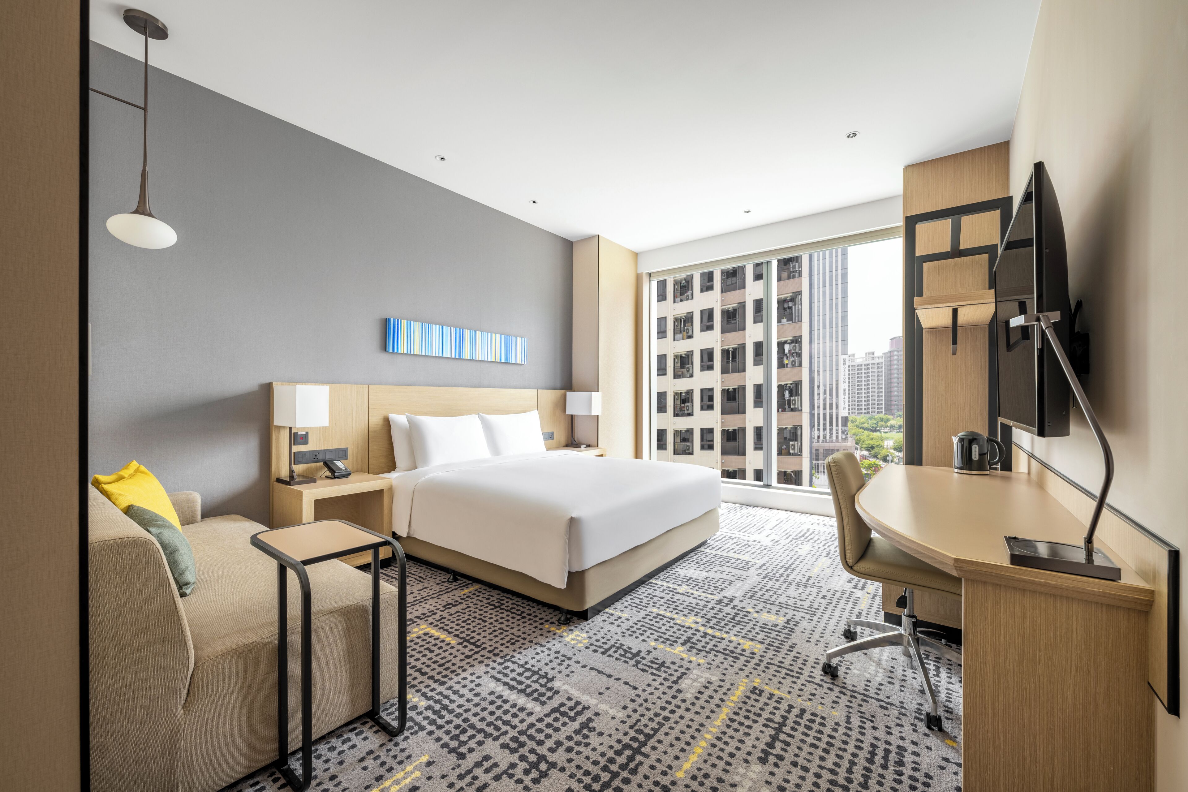 Habitación de hotel con cama, escritorio, silla y amplios ventanales con vista a la ciudad.
