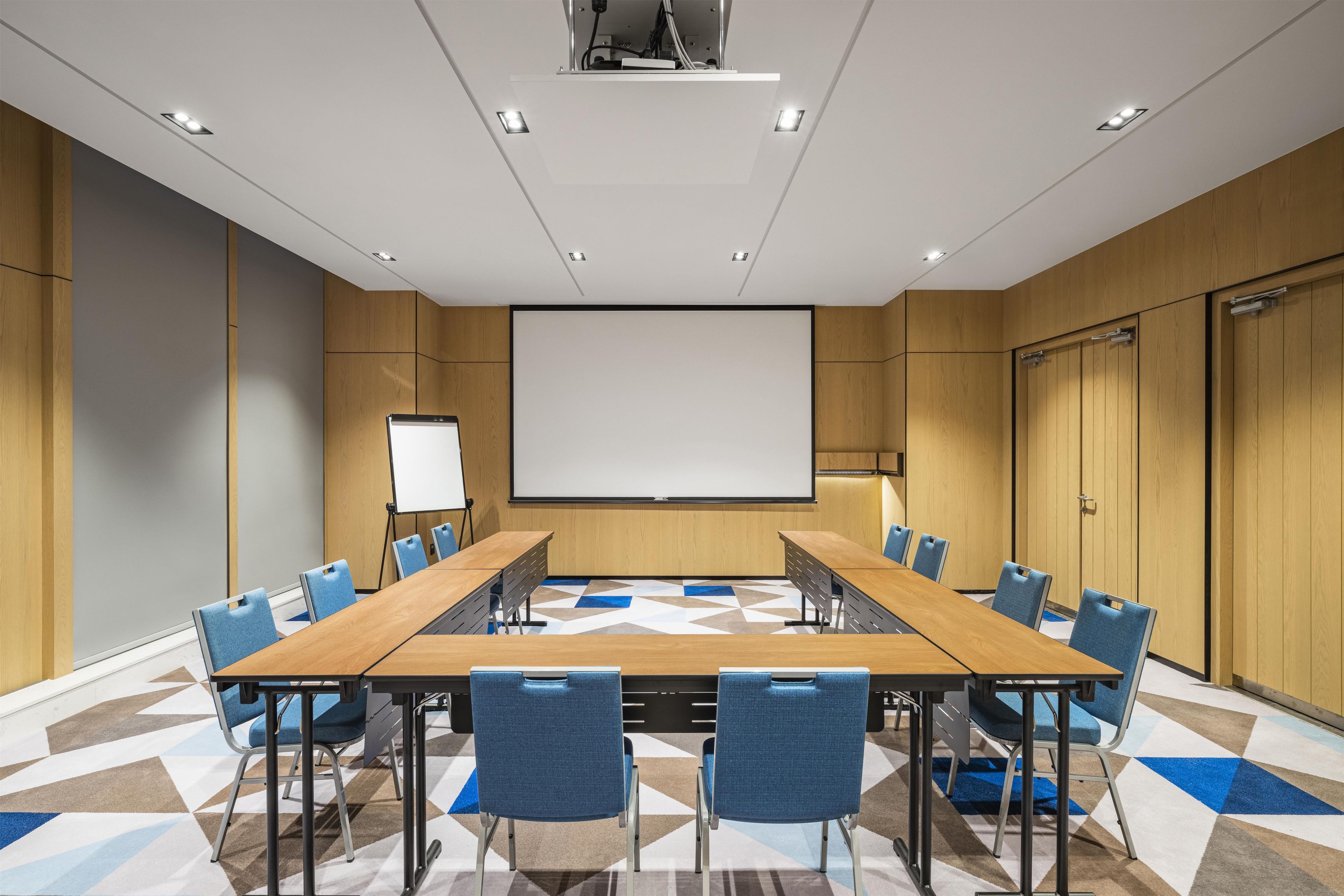 A conference room with a long table, blue chairs, a projector screen, and a whiteboard.