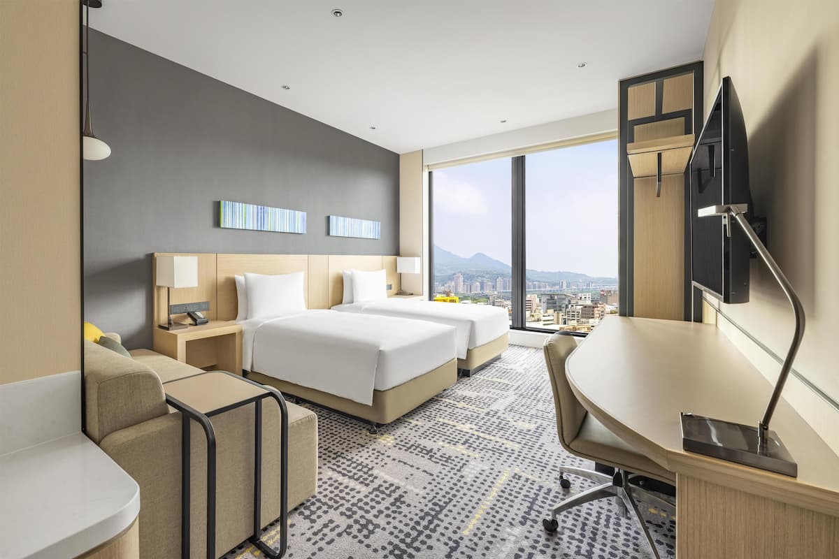 A hotel room with two beds, a desk, a chair, a TV, and a large window offering a city view.