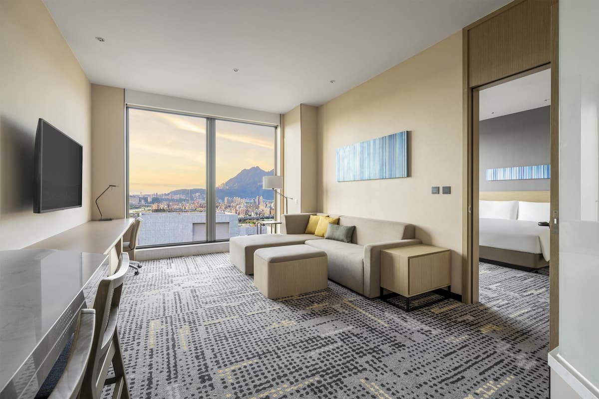 A modern hotel room with a large window offering a city view, a flat-screen TV, a sofa, a desk, and a bed.
