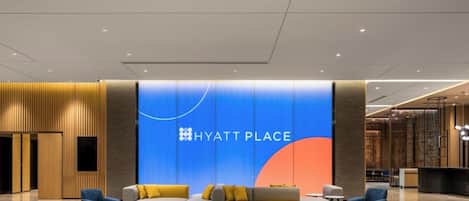 Un vestíbulo moderno con una gran pared azul que muestra el logotipo 'HYATT PLACE', un área de descanso con sofás y sillones, y un mostrador de recepción.