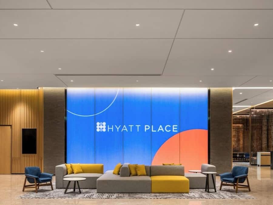 A modern lobby with a large blue wall displaying the 'HYATT PLACE' logo, a seating area with sofas and armchairs, and a reception desk.