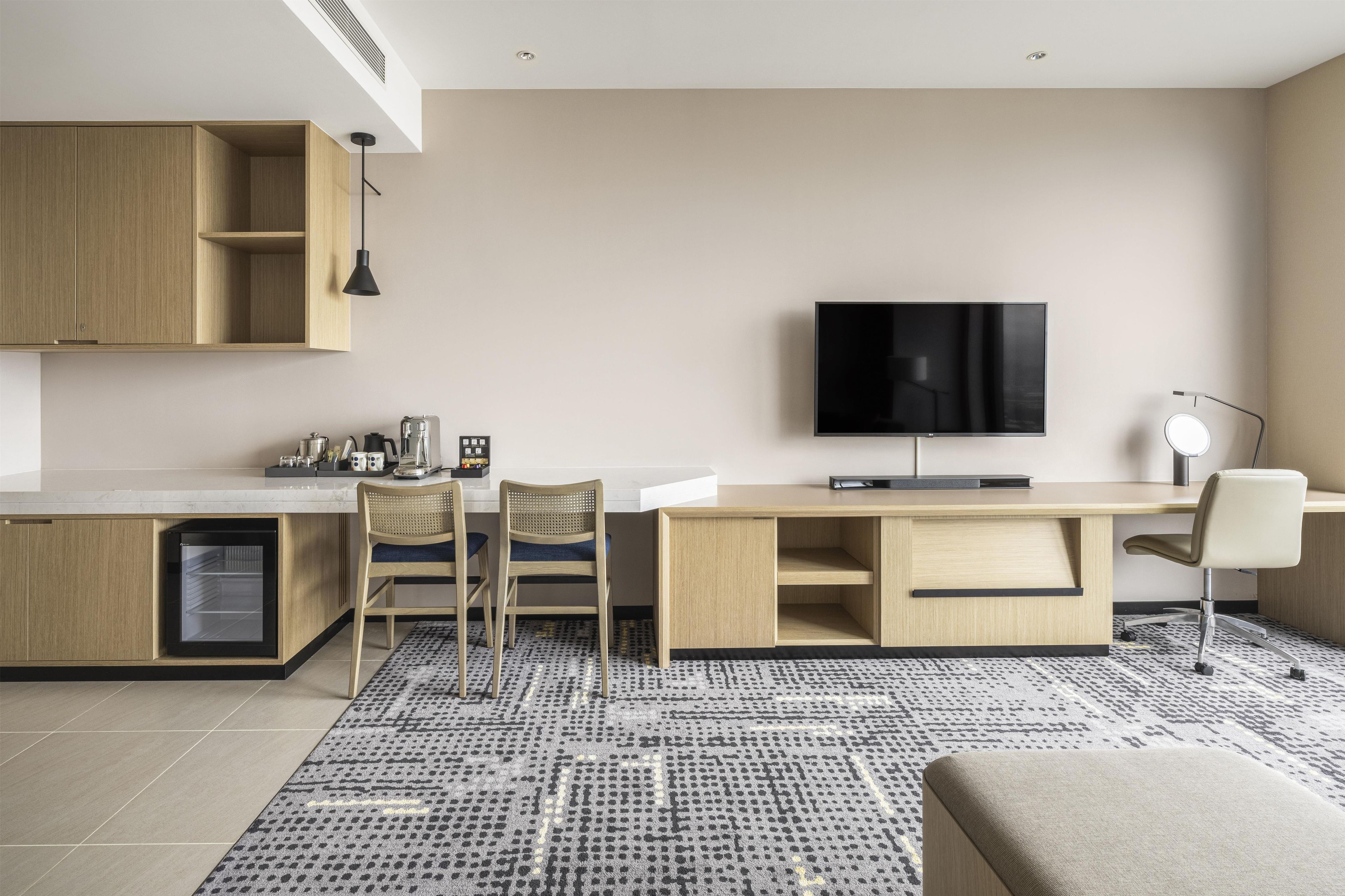 A modern kitchen with a built-in oven, a dining area with two chairs, a flat-screen TV mounted on the wall, and a small desk with a chair.