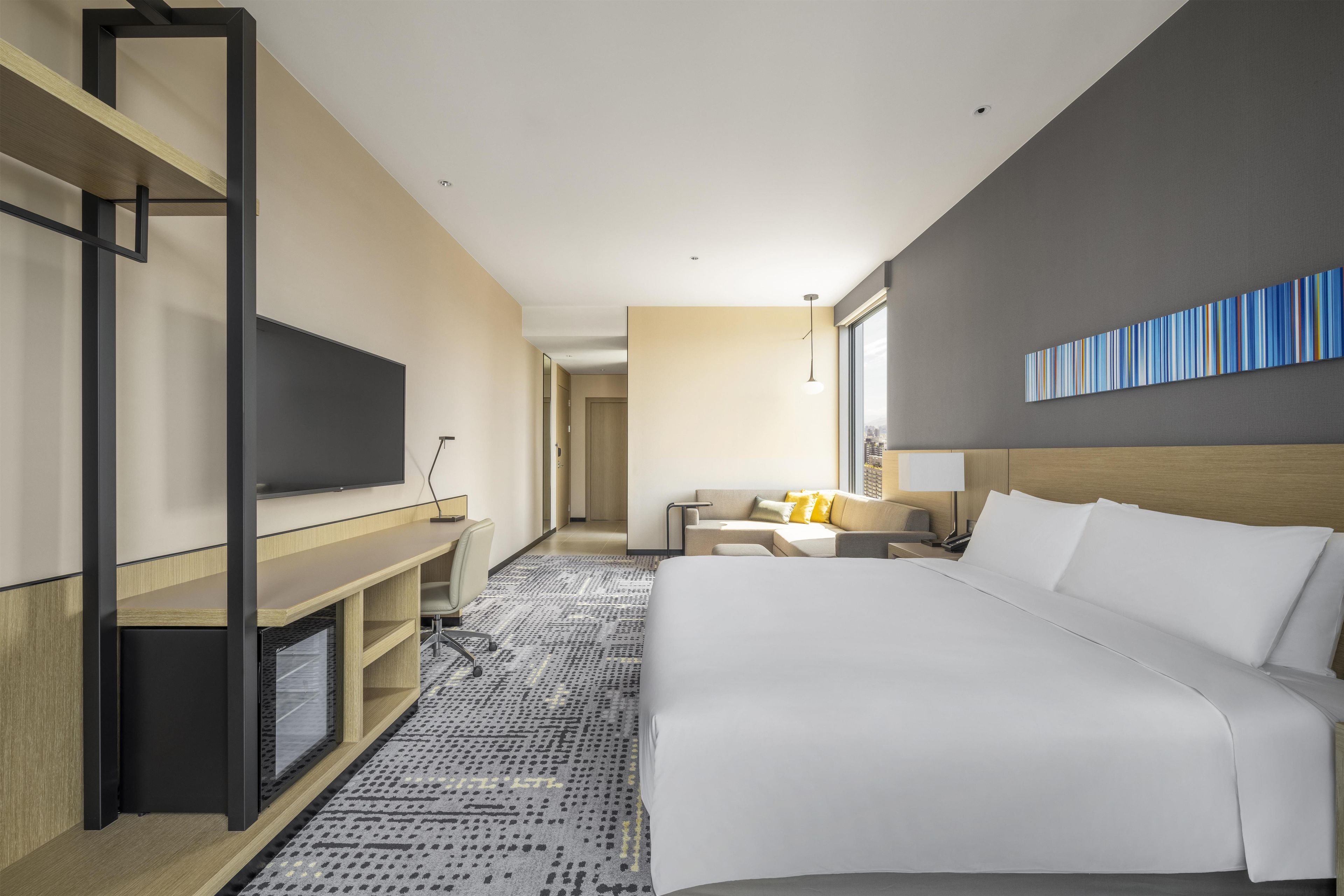 Quarto de hotel com uma cama grande, uma escrivaninha, televisão e vista para a cidade.