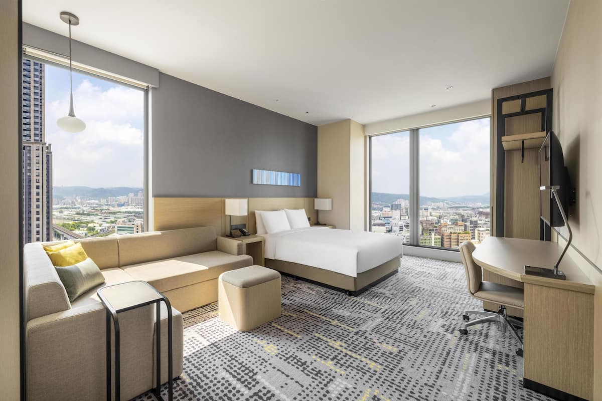 A modern hotel room with a large bed, a sofa, a desk, and a view of the city.