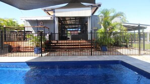 Uma piscina com área de estar coberta, cerca de metal e um edifício com telhado inclinado ao fundo.