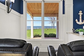 A room with a large window overlooking a grassy area and a body of water, two leather chairs, a wooden ceiling, and a white wall with an anchor decoration. - 12 Person Hot Tub Overlooking Lake Michigan (Pentwater)