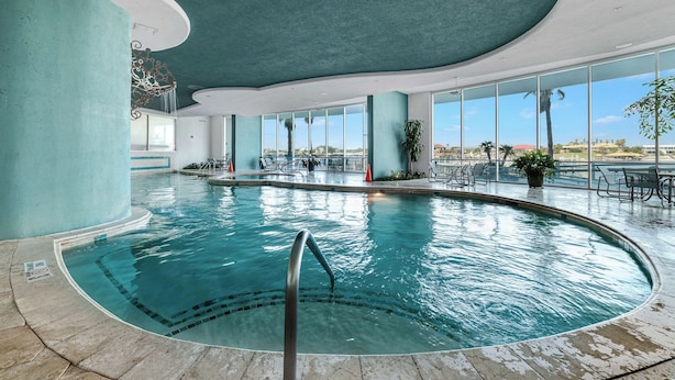 An indoor swimming pool with clear blue water, surrounded by large windows offering views of palm trees and buildings. - Bella Luna 1009-Luxury Condo-Great Views-Walk to Flora-Bama-Boaters Heaven! (Orange Beach)