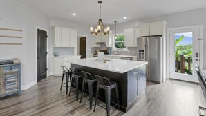 A modern kitchen with a large island, stainless steel appliances, and a view of greenery through a sliding door. - Serenity on Siverly: Saltwater pool and amazing view (McArthur)