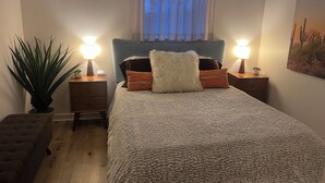 Une chambre à coucher comprenant un lit, deux tables de chevet, une petite table, une plante en pot et une fenêtre avec des rideaux.