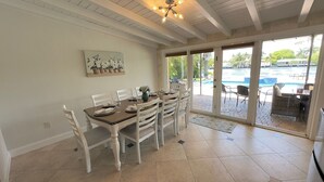 Un coin repas avec une table dressée pour quatre personnes, des chaises blanches, un ventilateur de plafond et une vue sur l’espace extérieur de la piscine.