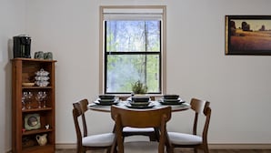A dining area with a round table set for four, surrounded by wooden chairs with cushioned seats. A wooden sideboard with glassware and decorative items is to the left. A window with a view of trees and a landscape painting is on the wall. - Deer Crossing Cabin Cabin in heart of Hochatown, 259, Broken Bow Lake (Broken Bow)