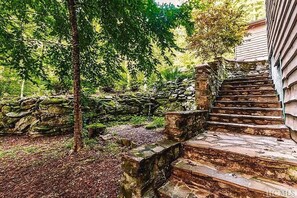 Un escalier en pierre mène à une maison en bois, entourée d’un jardin luxuriant et d’un mur de pierres.