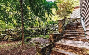 Eine Steintreppe führt zu einem Holzhaus, das von einem üppigen Garten und einer Steinmauer umgeben ist.