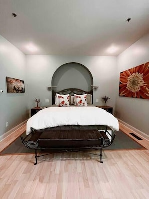 Un dormitorio con una cama centrada bajo un amplio techo abovedado, flanqueada por dos mesitas de noche con lámparas, un cuadro floral grande en la pared derecha y un cuadro abstracto más pequeño en la izquierda.