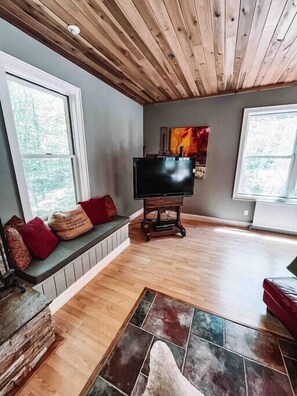 A living room with a wooden ceiling, a large flat-screen TV on a stand, a bench with cushions, and a stone tile floor.