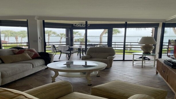 Ein geräumiges Wohnzimmer mit großem Fenster und Blick auf ein Gewässer, ausgestattet mit einer Couch, Sesseln und einem Couchtisch.