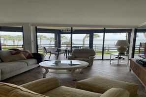 A spacious living room with a large window overlooking a body of water, furnished with a sofa, armchairs, and a coffee table.