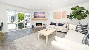 A spacious living room with a large sofa, a coffee table, and a fireplace. There is a large window with a view of the outside. - 3 Bedroom Renovated Condo on Balboa Island (Newport Beach)