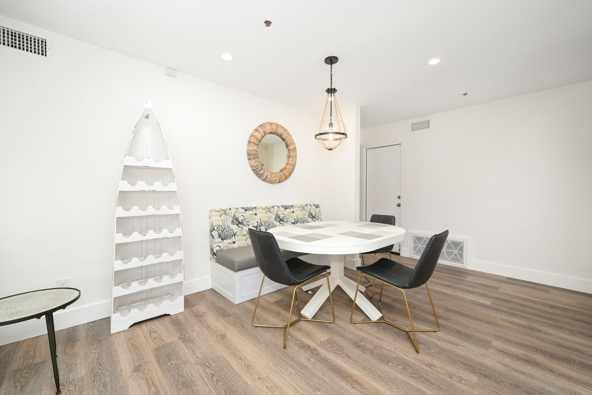 A dining area with a round table, black chairs, a white bench, a decorative wall piece, and a hanging light fixture.