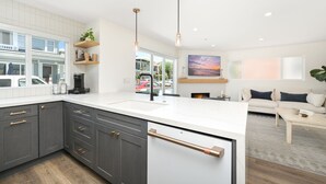 A modern kitchen with dark grey cabinets, a white countertop, and a stainless steel range hood. There is a dining area with a white table and a sofa. - 3 Bedroom Renovated Condo on Balboa Island (Newport Beach)