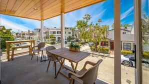 A covered patio with a wooden table and chairs, large windows offering a view of a residential area with houses and palm trees.