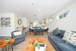 A modern living room with a grey sofa, a coffee table with a plant, and a dining area with blue chairs and a wooden table. - 4 bedroom Renovated Home on Balboa Island (Newport Beach)