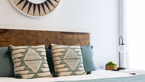 Un lit bien fait, agrémenté d’oreillers à motifs, une tête de lit en bois, une table de chevet avec une plante et une horloge murale.