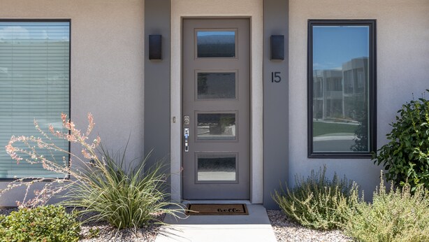 Une maison moderne avec une porte grise, le numéro 15, et un petit jardin agrémenté de plantes et d’un paillasson.