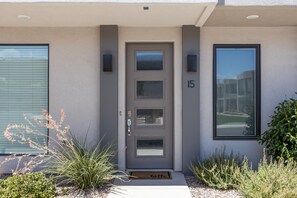 Ein modernes Haus mit grauer Tür, Hausnummer 15, einem kleinen Garten mit Pflanzen und einer Fußmatte.