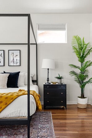 A four-poster bed with a white and mustard blanket, a black nightstand with a lamp, a potted plant, and a window with blinds.