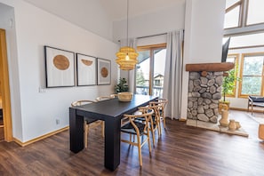 A dining area with a long table, wooden chairs, a hanging light fixture, and framed artwork on the wall.