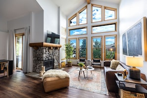 A spacious living room with a stone fireplace, large windows, and a cozy seating area. - All Seasons Retreat- 2BR+Loft 3BA-Upscale Design, Gorgeous Open Spaces (Mammoth Lakes)
