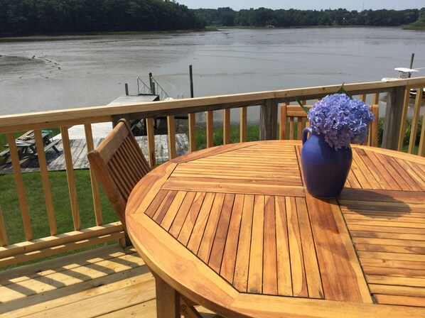 A wooden deck with a round table and chairs, overlooking a river with boats.
