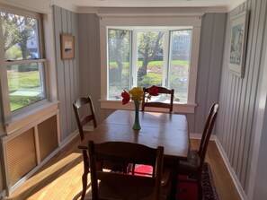 A dining area with a wooden table and chairs, a vase with flowers, and large windows with a view of a yard and trees.