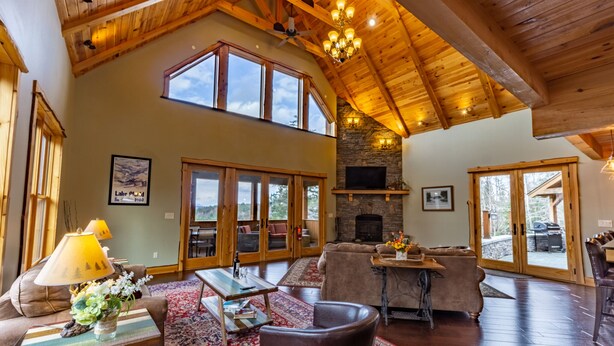 A spacious living room with a high ceiling, wooden beams, a stone fireplace, and large windows. - Stunning Mountaintop Retreat with High End Amenities and Four Fireplaces (North Creek)