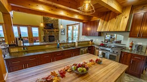 A spacious kitchen with wooden cabinets, a large island with a bowl of fruit, and a stone fireplace. - Stunning Mountaintop Retreat with High End Amenities and Four Fireplaces (North Creek)