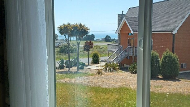 A view through a window of a building with a staircase leading to a porch, a lawn, and a distant view of the ocean. - Great location on a 9 hole, golf course, short walk to the ocean. (Shelter Cove)