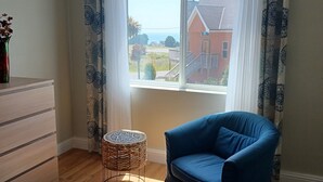 A room with a large window, a blue chair, a wooden dresser, and a wicker stool. - Great location on a 9 hole, golf course, short walk to the ocean. (Shelter Cove)