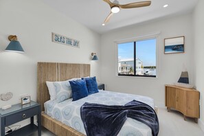 A bedroom with a bed, a nightstand, a wooden cabinet, a ceiling fan, and a window with a view of buildings and boats. - Catch n' Relax Anglers Ultimate Waterfront Retreat, 50’ Dock, Pool Home Modern (Marathon)