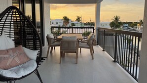 A balcony with a table and chairs, a wicker chair, and a view of a sunset over a marina.