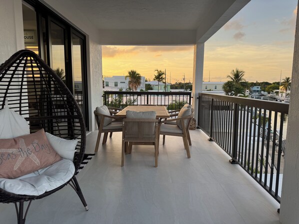A balcony with a table and chairs, a wicker chair, and a view of a sunset over a marina.