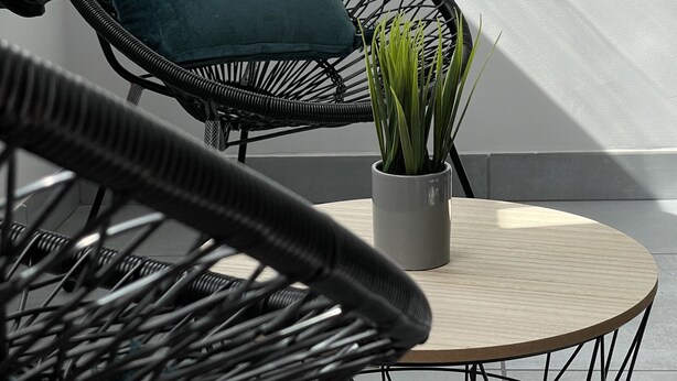 Una silla de mimbre con un cojín azul turquesa, una planta pequeña en maceta y una mesa redonda.