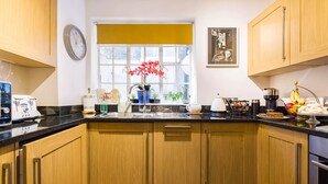 A kitchen with wooden cabinets, a black countertop, a window with a yellow blind, a clock, and various kitchen appliances and items. - Mayfair Centre 3-Bed 6-Guest Art Nouveau Flat in 1729 Private House (London)