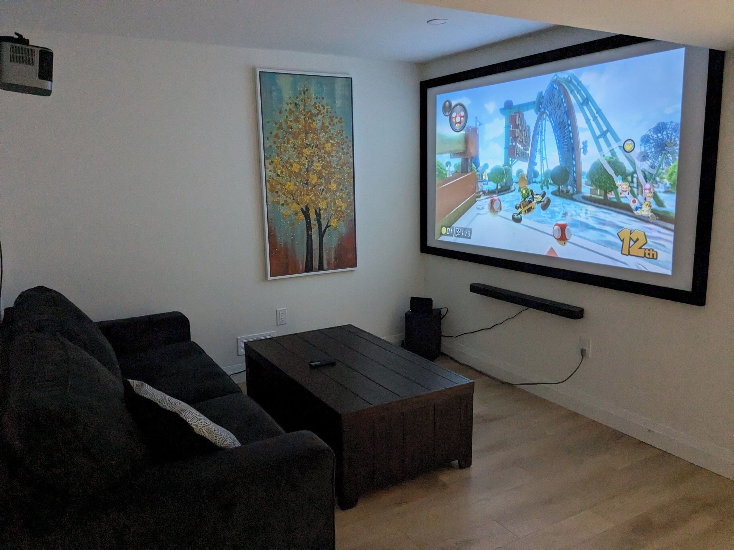 Un salon avec un grand écran diffusant un jeu vidéo, un canapé, une table basse et un tableau accroché au mur.