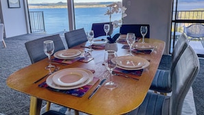 Une table à manger dressée avec des assiettes, des verres et des couverts, offrant une vue panoramique sur la mer et le paysage lointain.