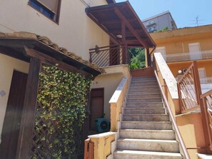 Una scala conduce a un balcone con ringhiera in legno, una porta con stipite in legno e un muro ricoperto di piante rampicanti.